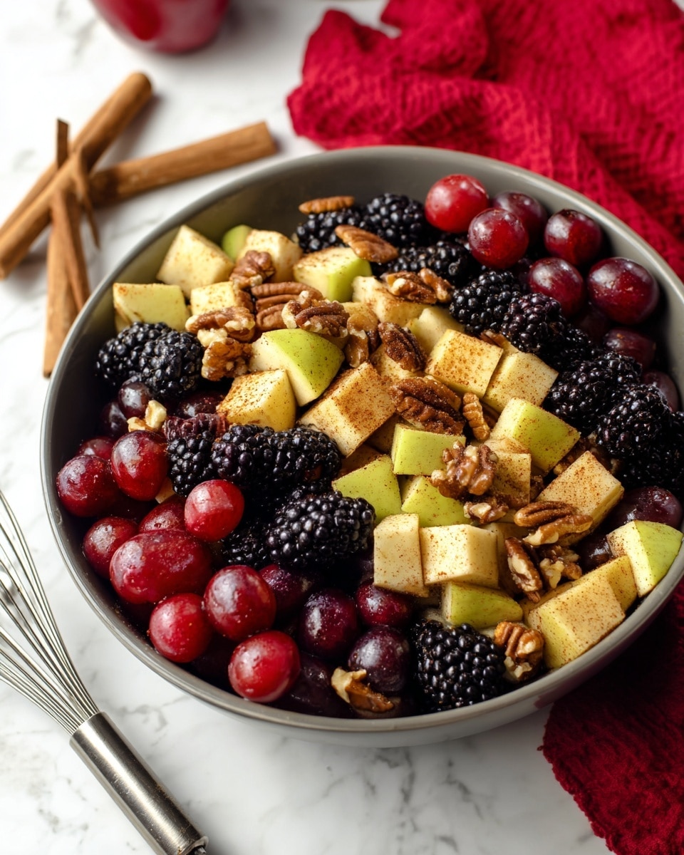A white bowl filled with a fruit salad showing three layers: the bottom layer with shiny red grapes, the middle layer with blackberries, and the top layer with light yellow apple cubes sprinkled with brown cinnamon powder. Mixed throughout are pecan halves adding a brown crunchy texture. The bowl is on a white marbled surface next to two cinnamon sticks and a metal whisk, with a red patterned cloth in the background. photo taken with an iphone --ar 4:5 --v 7