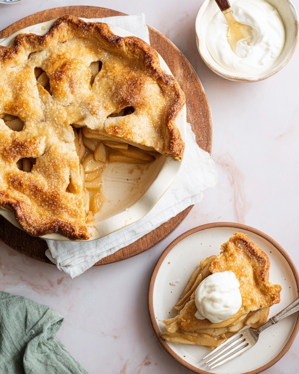 A round apple pie with a golden brown, slightly cracked top crust sits in a white pie dish, with one large slice missing, showing soft, cooked apple slices layered inside. The crust looks flaky and sprinkled with sugar crystals. A single slice is placed on a white plate beside the pie, revealing the light brown apple filling under a golden crust, topped with a dollop of white whipped cream. A fork with a wooden handle rests on the plate next to the slice. Nearby, there is a small white bowl filled with more whipped cream. All items are set on a white marbled surface. Photo taken with an iphone --ar 4:5 --v 7