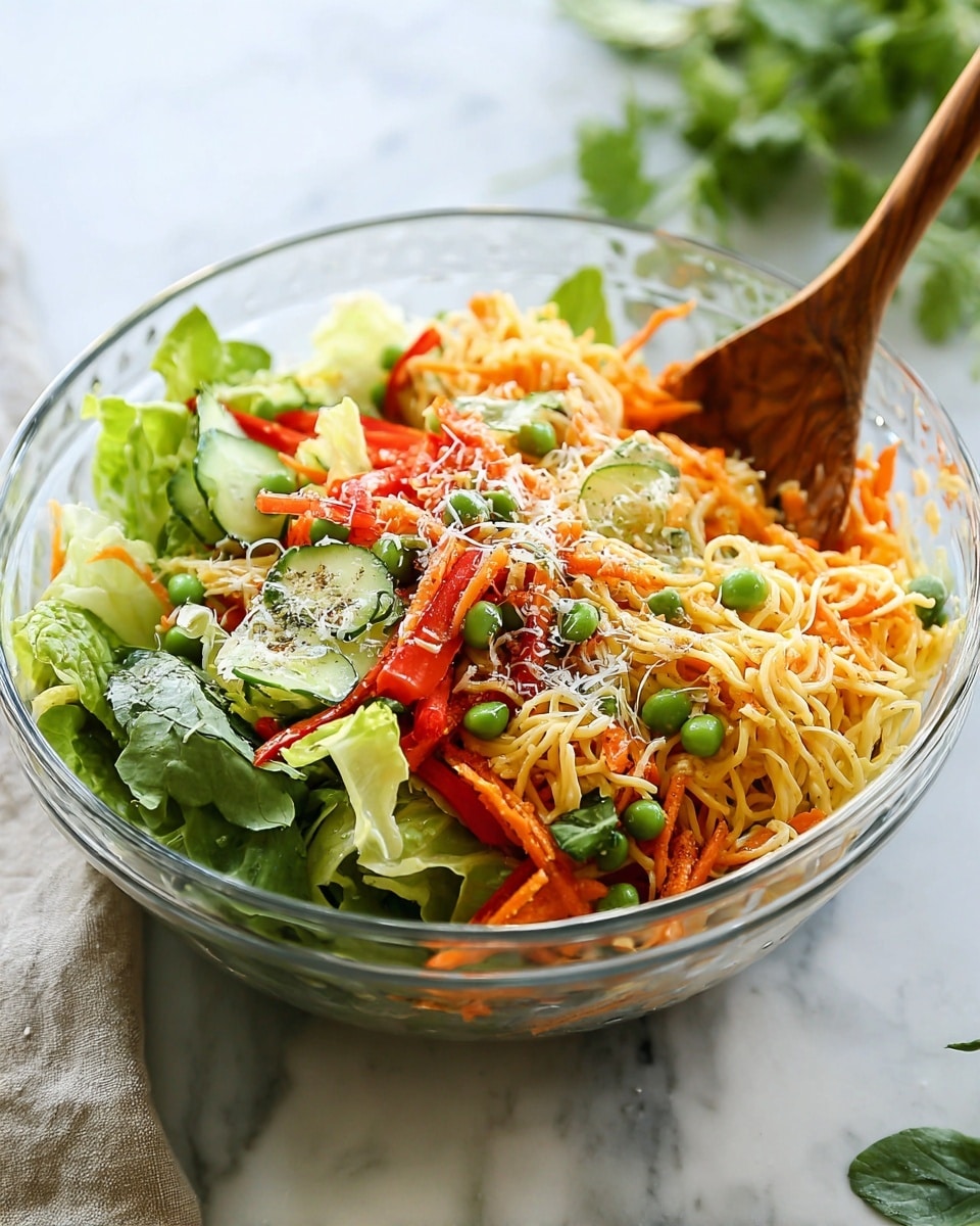 A clear glass bowl holds a colorful mixed salad with multiple layers on a white marbled surface. The base layer features light cream-colored thin noodles, topped with green leafy lettuce, bright orange shredded carrots, and thin slices of green cucumber. On top of these are deep red strips of bell pepper, scattered green peas, and a light sprinkle of grated white cheese. A wooden spoon is placed inside the bowl on the right side. In the background, some blurred green herbs add a fresh touch. A white cloth is visible on the left near the bowl. Photo taken with an iphone --ar 4:5 --v 7