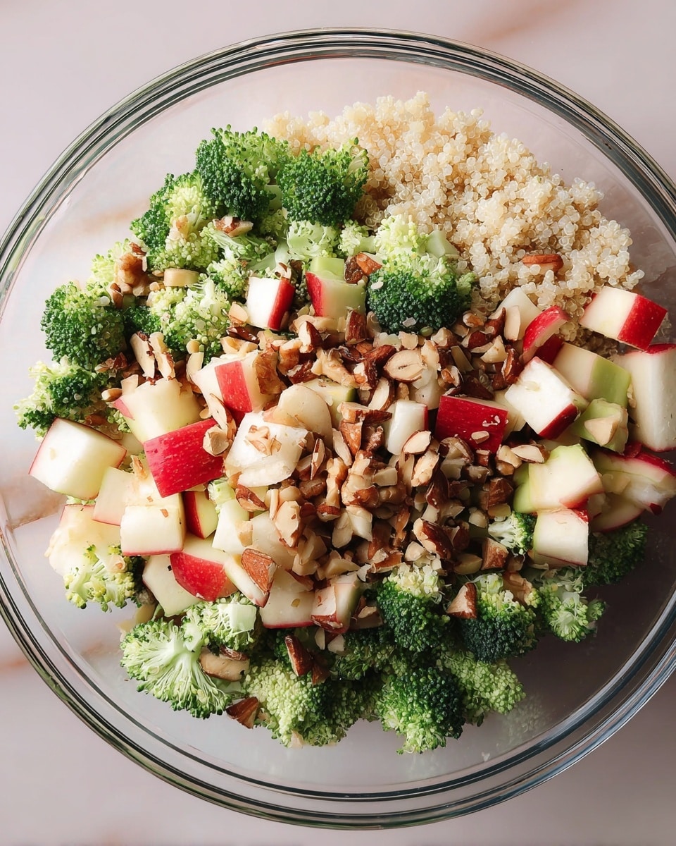 The image shows a clear glass bowl filled with a colorful salad from a top view. The salad has several layers mixed together: small green broccoli florets, light beige quinoa grains, white crumbled cheese, red and white chopped apple pieces, and light brown chopped walnuts. All ingredients are evenly spread and mixed, giving the salad a fresh and healthy look. The bowl is placed on a white marbled surface. photo taken with an iphone --ar 4:5 --v 7