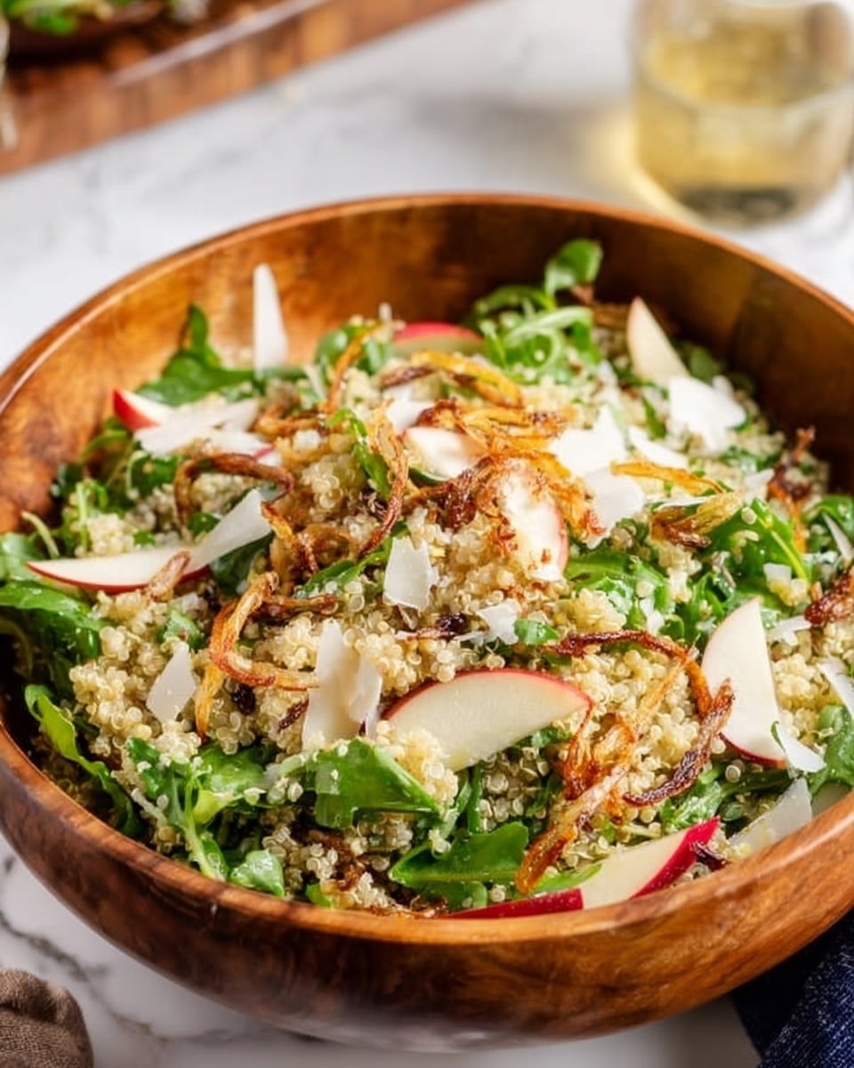 A wooden bowl filled with a fresh quinoa salad shows several layers: at the base, light beige quinoa grains mixed with green leafy arugula, thin slices of red apple with white flesh spread evenly throughout, topped with small white cheese shreds and crisp golden-brown fried shallots scattered around. The colors contrast nicely, with the light grains and pale apple slices balancing the green leaves and browned shallots. The bowl sits on a white marbled surface, with a glimpse of a yellow dressing in a small glass bowl in the background. photo taken with an iphone --ar 4:5 --v 7