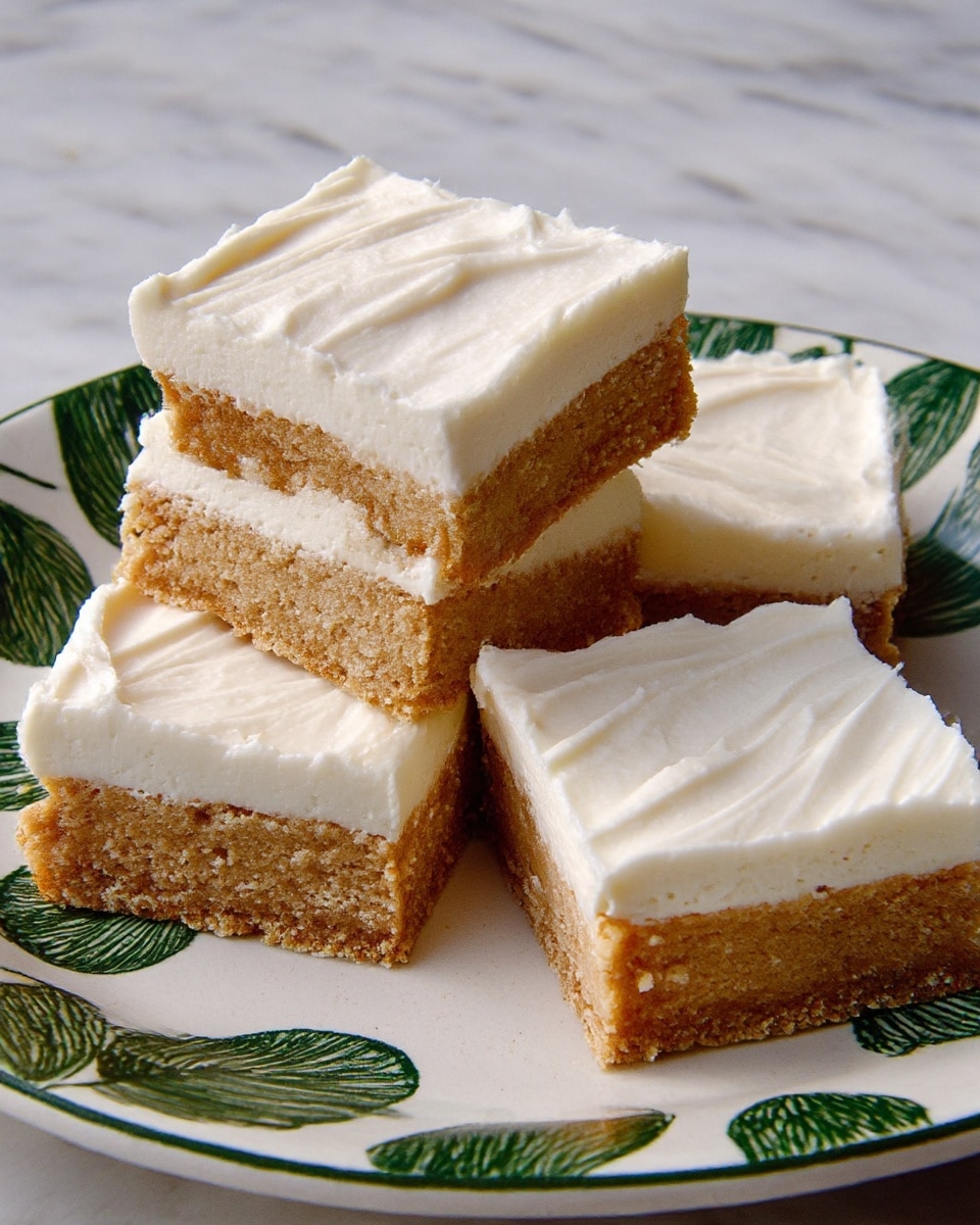 Four square pieces of dessert are shown on a white plate with green leaf patterns. Each piece has two layers: the bottom layer is thick and light brown with a soft, crumbly texture, while the top layer is a thick, creamy white frosting with smooth swirls and a slightly fluffy look. One piece is stacked on top of another, slightly lifted to show the clear separation between the brown base and white frosting. The surface under the plate is a white marbled texture. photo taken with an iphone --ar 4:5 --v 7