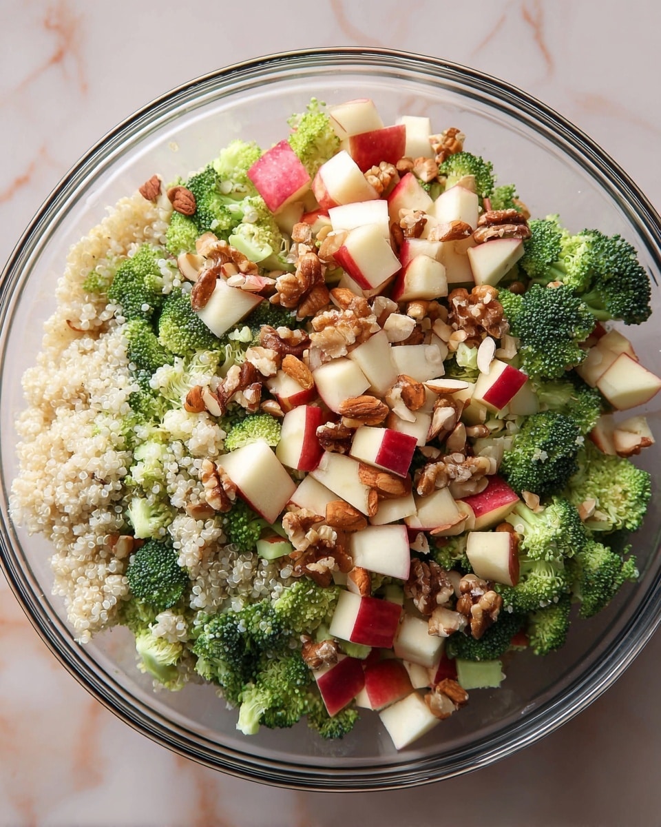 A clear glass bowl filled with a colorful salad showing multiple layers: the bottom layer has fluffy white quinoa grains, followed by bright green broccoli florets scattered evenly, topped with small cubes of red-skinned apple mixed with chopped brown walnuts and light beige almond slices, all sitting on a white marbled surface. photo taken with an iphone --ar 4:5 --v 7