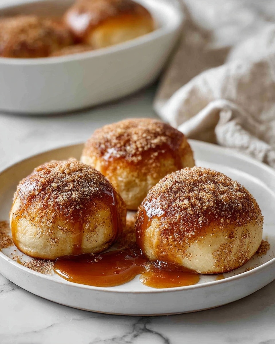 Three soft, round buns sit closely on a white plate with a white marbled texture underneath. Each bun is golden brown with a slightly crisp surface and is thickly coated on top with a grainy mixture of cinnamon and sugar dusted generously. Rich caramel sauce drips slowly down the sides and pools slightly on the plate, giving the buns a shiny, sticky look. In the background, a white dish holds more buns, softened by the out-of-focus effect, and a slightly crumpled off-white cloth lies nearby. The whole scene is warm and inviting with natural lighting. photo taken with an iphone --ar 4:5 --v 7