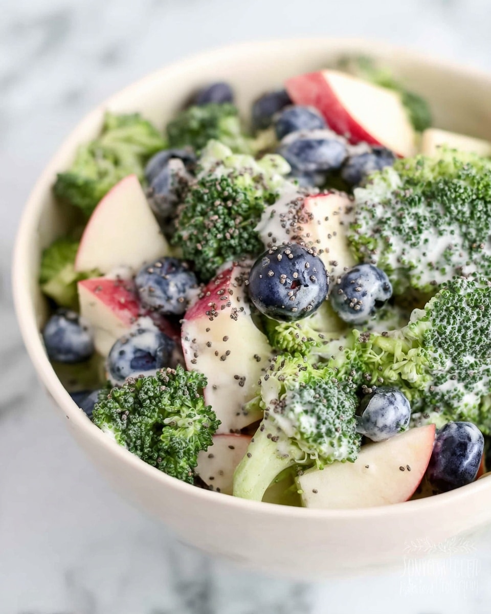 A white bowl filled with a fresh salad that has three main layers: small bright green broccoli florets with a slightly rough texture at the base, medium-sized round blueberries with a smooth deep purple surface scattered throughout the salad, and small dice pieces of red-skinned apple with cream-colored flesh. Everything is mixed and lightly covered with creamy dressing that has tiny black poppy seeds spread evenly on top. The bowl is placed on a white marbled surface. photo taken with an iphone --ar 4:5 --v 7