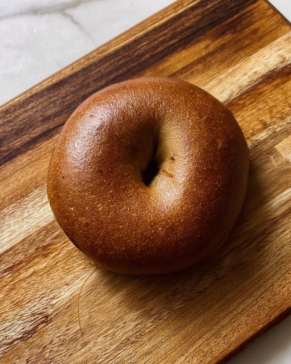 A single round bagel with a smooth, shiny, and golden-brown crust sits on a wooden cutting board with visible wood grain and natural color variations. The bagel has a slightly wrinkled texture and a small hole in the center, showing classic bagel shape and density. The wooden surface contrasts with the warm, rich color of the bagel. The background is a white marbled texture. Photo taken with an iphone --ar 4:5 --v 7