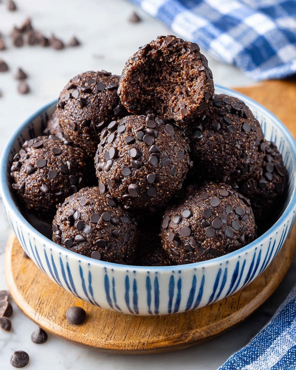 A white bowl with blue vertical stripes is filled with round chocolate cookie dough balls covered in small dark chocolate chips. The cookie dough balls are dark brown with a rough texture and uneven surface, each densely coated with shiny dark chocolate chips that add a speckled look. One ball is bitten, showing a soft and crumbly inner texture with a slightly grainy appearance. The bowl sits on a wooden board with scattered chocolate chips around it, and a blue and white checkered cloth is partly visible in the background. The scene is set on a white marbled texture. photo taken with an iphone --ar 4:5 --v 7