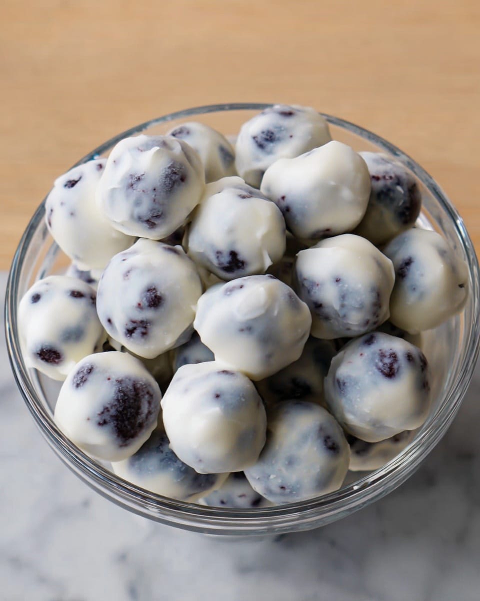 The image shows a bowl filled with many small round treats, each covered in a smooth, white coating with a slightly glossy texture. Under the white coating, hints of dark purple or black spots can be seen, suggesting the treats are made with blueberries or a similar fruit. The bowl is transparent and placed on a white marbled surface. The treats are piled closely together, filling the bowl almost to the top, creating a soft, rounded texture overall. photo taken with an iphone --ar 4:5 --v 7