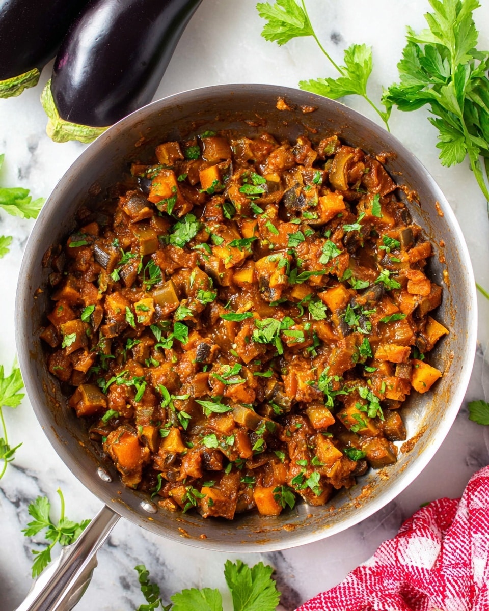 A silver pan filled with a cooked dish made of small, evenly cut dark orange and brown cubes, coated in a thick brown sauce, topped with fresh green chopped parsley scattered across. The pan sits on a white marbled surface, surrounded by whole dark purple and green eggplants and some flat green parsley leaves. A part of a red and white checkered cloth is visible near the bottom right corner. Photo taken with an iphone --ar 4:5 --v 7