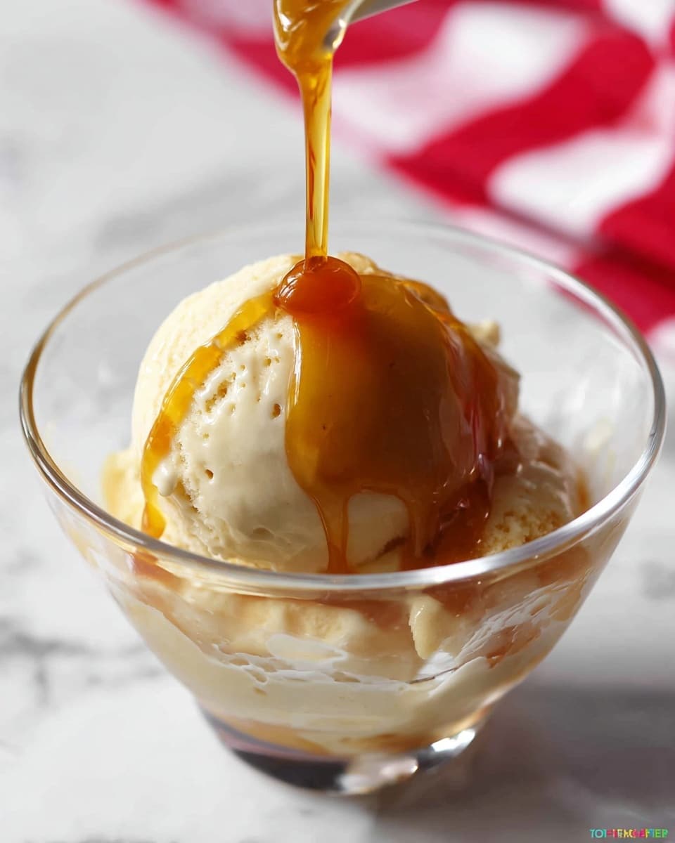 A clear glass bowl holds a single scoop of creamy white ice cream with a smooth texture and visible small air pockets. A golden brown liquid is being poured slowly over the ice cream from above, creating a glossy, thick layer that drips down the sides. The bowl sits on a white marbled surface, with a blurred red and white cloth in the background. photo taken with an iphone --ar 4:5 --v 7
