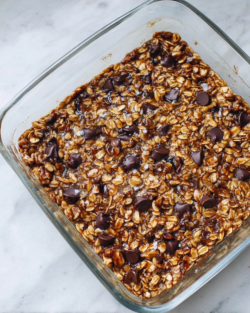 The image shows a single-layer baked oatmeal dish in a clear glass square baking dish. The top layer is covered with toasted golden brown oats mixed with visible dark brown walnuts and scattered dark chocolate chips melted slightly, adding a glossy texture. There are also small flakes of sea salt sprinkled on top, contrasting with the warm, earthy colors. The dish sits on a white marbled surface. photo taken with an iphone --ar 4:5 --v 7