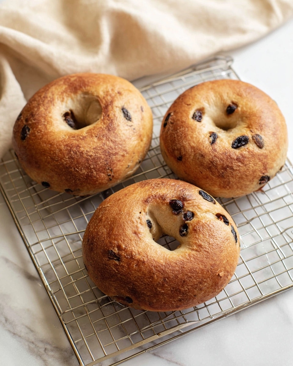 Three golden brown bagels with dark spots from raisins or olives are placed on a silver metal cooling rack. Each bagel has a round shape with a hole in the middle, and the surfaces look slightly shiny and textured with some small cracks. The rack sits on a smooth white marbled surface, and in the background, there's a soft beige cloth adding a cozy feel. Photo taken with an iphone --ar 4:5 --v 7