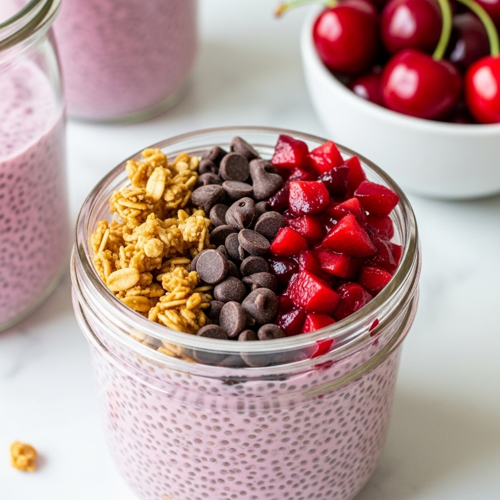 The image shows a glass jar filled with three layers: the bottom and main layer is creamy pink chia pudding with a textured surface full of chia seeds. On top, there are three different toppings arranged side by side: light brown crunchy granola on the left, smooth and dark brown chocolate chips in the middle, and shiny, deep red diced cherries on the right. In the background, there is another glass jar filled with more pink chia pudding, and a white bowl with whole red cherries sitting on a white marbled surface. Photo taken with an iphone --ar 4:5 --v 7