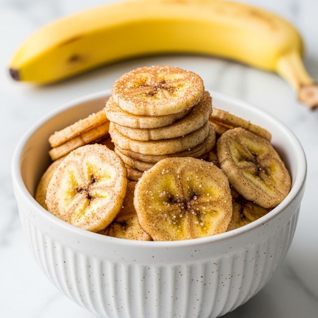 A white bowl filled with many banana chips stacked on top of each other, each chip golden yellow with a light crispy texture and dusted evenly with brown cinnamon and sugar granules, creating a sparkling effect. The bowl has vertical ridges and small speckles of brown. In the blurred background, a whole banana sits on a white marbled surface. photo taken with an iphone --ar 4:5 --v 7