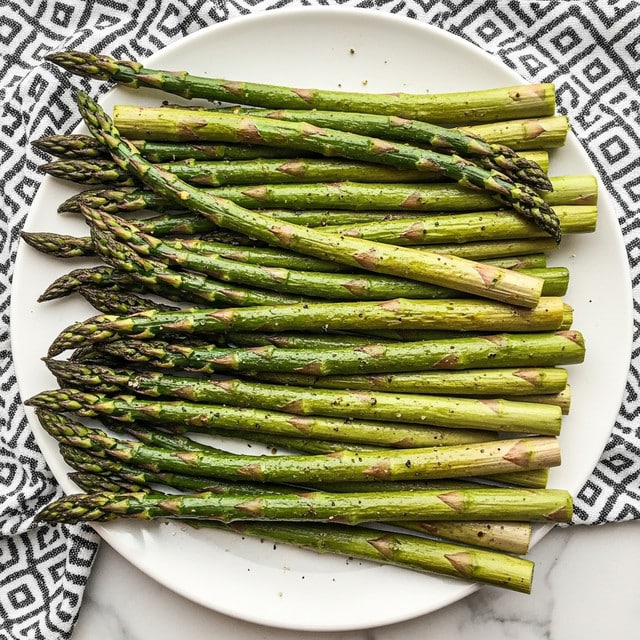 The image shows a white plate full of cooked asparagus stalks, arranged in a neat pile. The asparagus is bright green with some charred and roasted spots, showing a slightly wrinkled texture and hints of seasoning like salt and pepper sprinkled on top. The stalks are long and slender, overlapping each other with the tips pointing in different directions. The plate sits on a white marbled surface with a black and white patterned cloth partially visible to the side. photo taken with an iphone --ar 4:5 --v 7